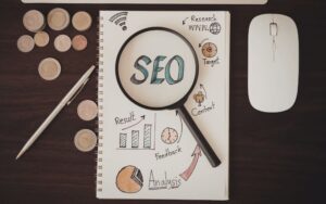 SEO in blauen Buchstaben auf einem weißen Zettel mit einer Lupe und Grafiken, umgeben von einem Stift, Münzen und einer Maus