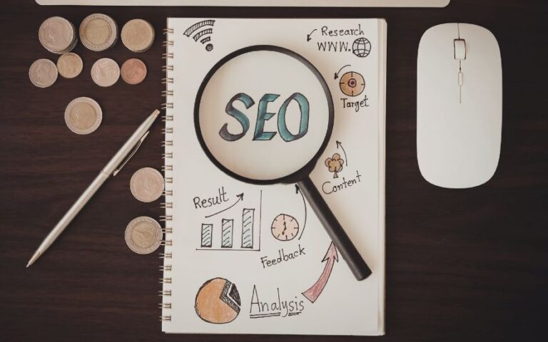 SEO in blauen Buchstaben auf einem weißen Zettel mit einer Lupe und Grafiken, umgeben von einem Stift, Münzen und einer Maus