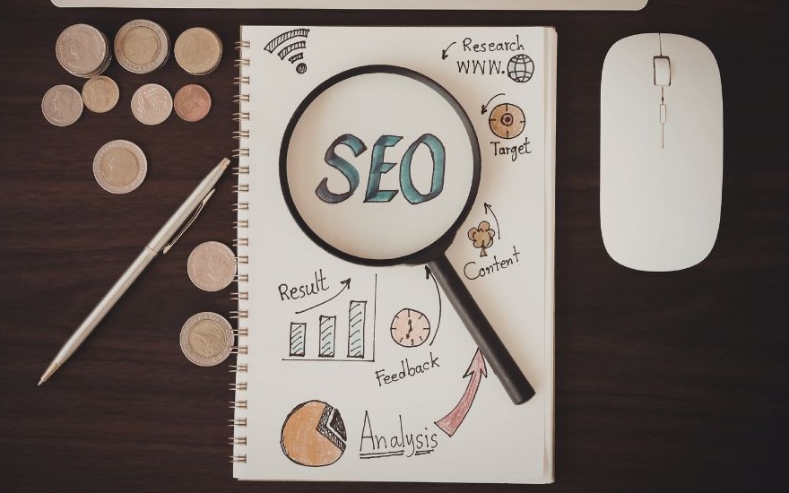 SEO in blauen Buchstaben auf einem weißen Zettel mit einer Lupe und Grafiken, umgeben von einem Stift, Münzen und einer Maus