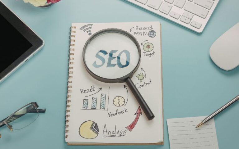 SEO in blauen Buchstaben auf einem weißen Zettel mit einer Lupe umgeben von einer Tastatur, Laptop, Brille, Stift und Maus